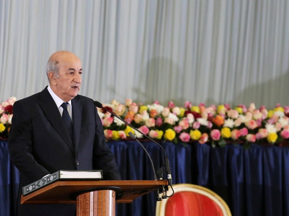 Homme âgé parlant à une conférence avec décor floral en arrière-plan.