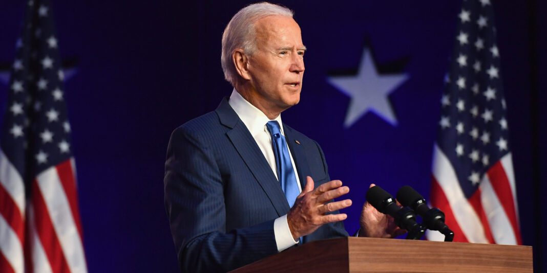 Joe Biden parle lors d'une conférence avec drapeaux américains en arrière-plan.
