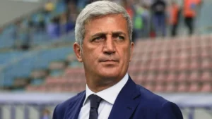 Homme en costume en stade, regard sérieux, contexte sportif.