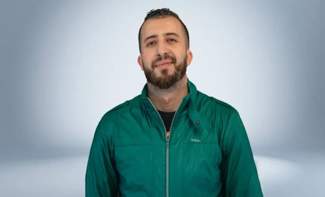 Anes Tina souriant, portant une veste verte, dans un studio neutre.