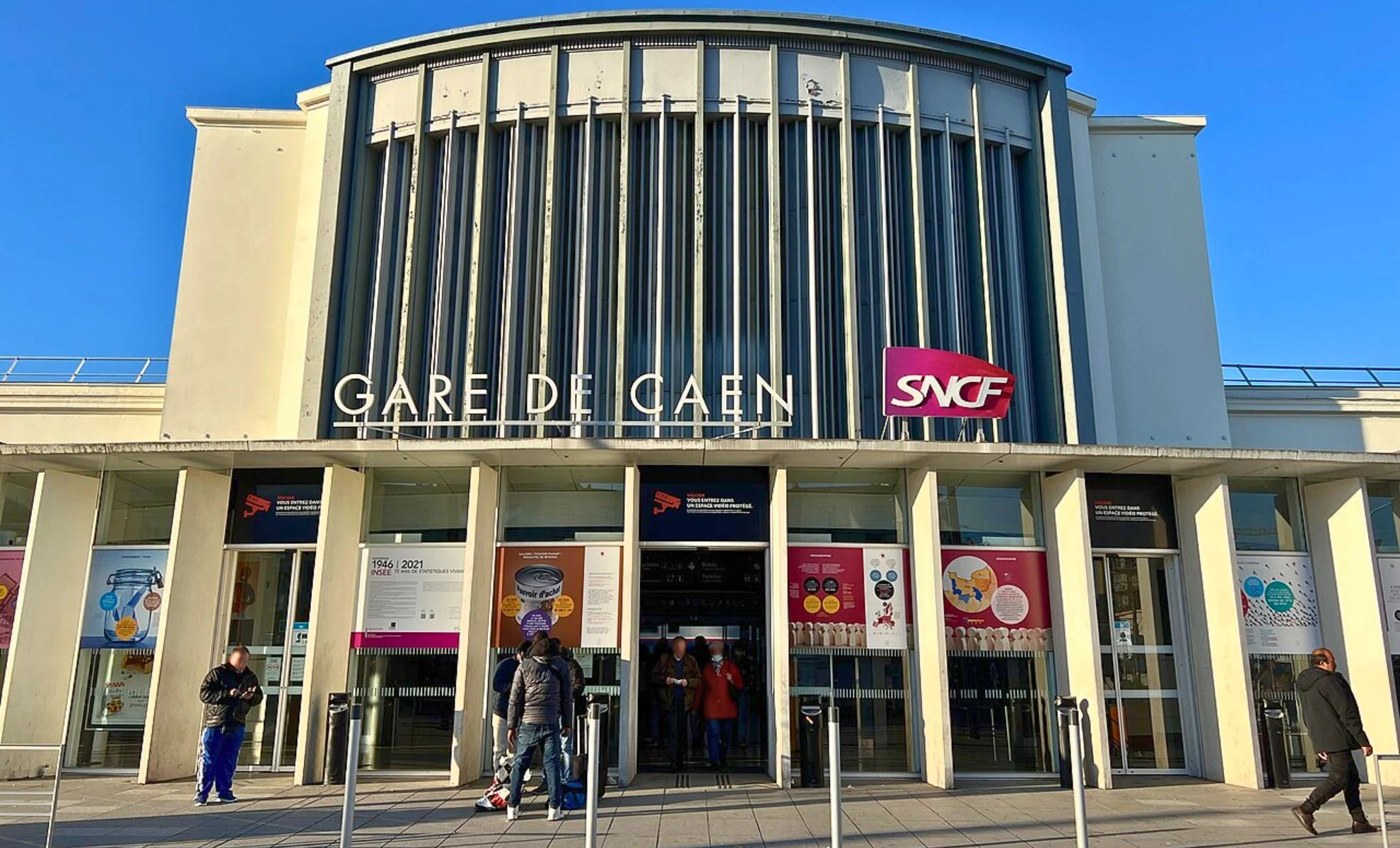 Façade de la gare de Caen avec logo SNCF et voyageurs.
