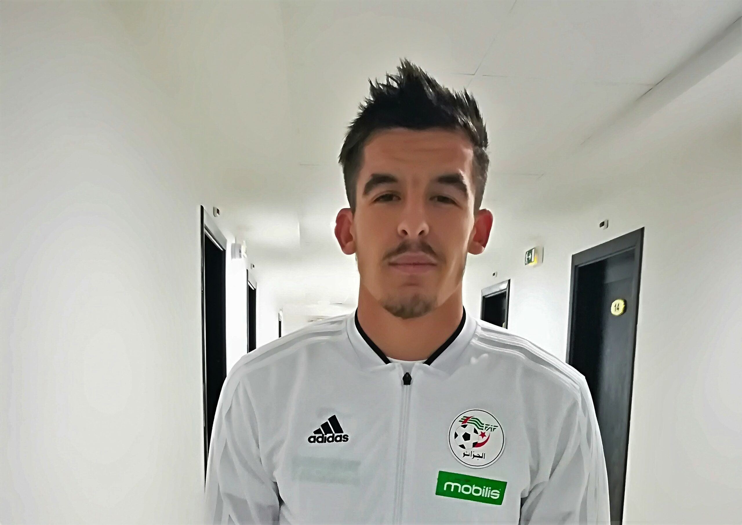 Gaya Merbah dans un couloir, expression sérieuse, en tenue de sport blanche.