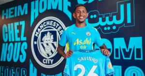 Savinho avec maillot Manchester City, sourire, fond logo club et mots en arabe et français.