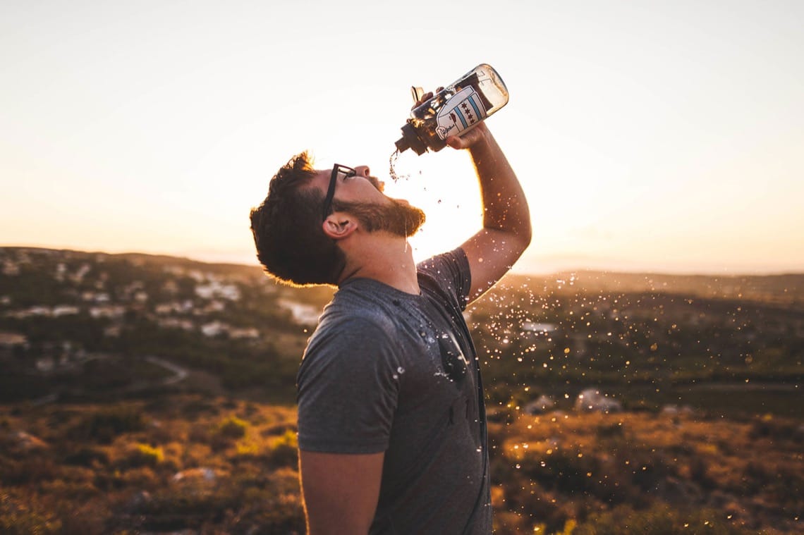 Homme portant des lunettes buvant une boisson en extérieur au coucher du soleil.