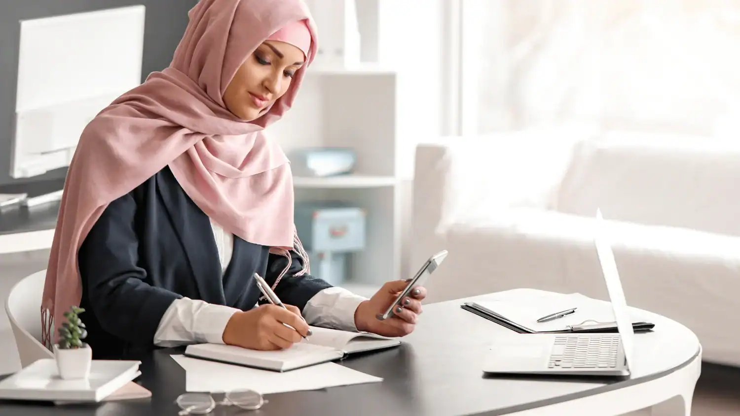 France : quand le voile coûte leur emploi aux salariées Femme avec hijab utilisant un smartphone et prenant des notes au bureau.