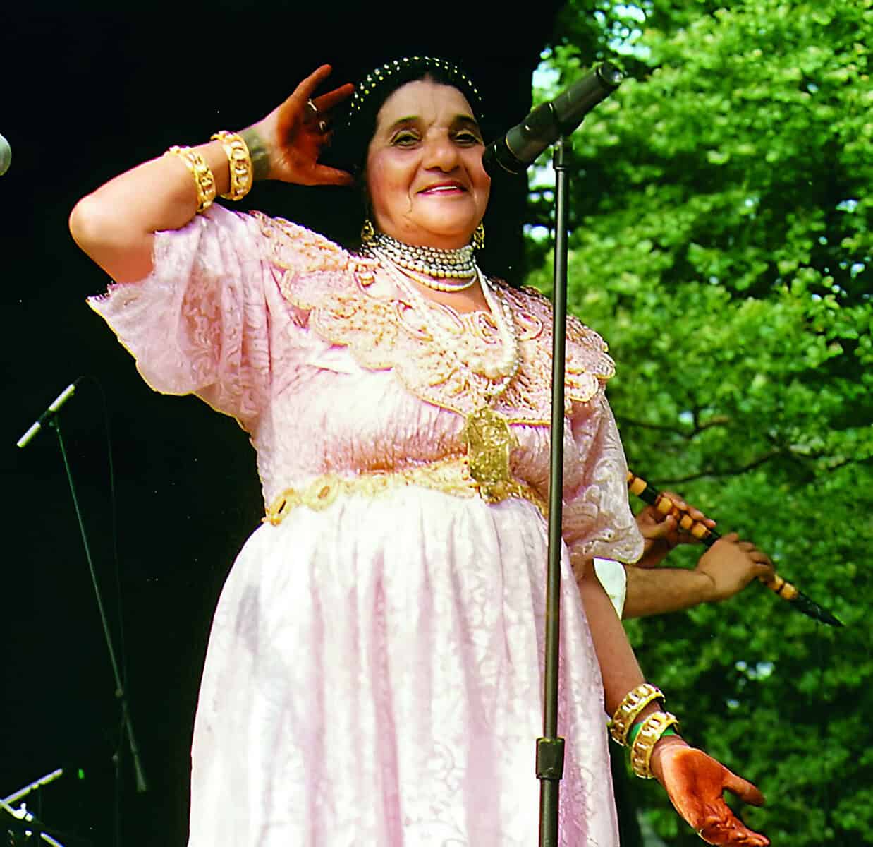 Cheikha Rimitti en performance, vêtue d'une robe rose et bijoux, chantant sur scène en plein air.