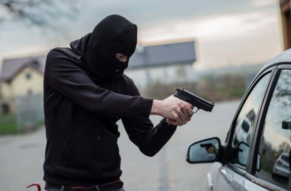 Homme masqué armé pointant un pistolet vers une voiture.