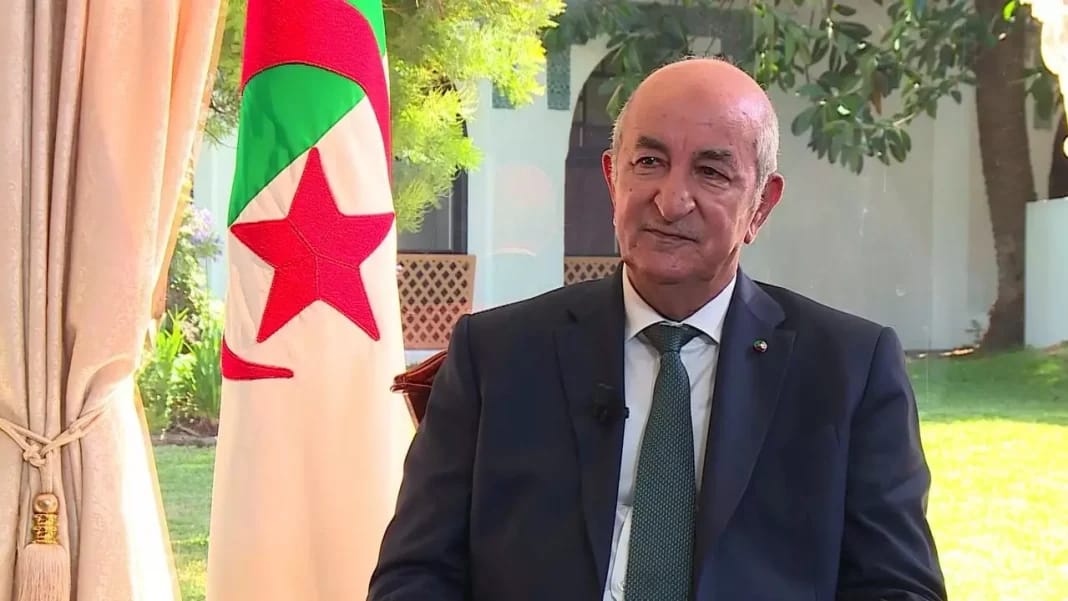 Réunion du Gouvernement : plusieurs secteurs à l’ordre du jour Homme politique algérien en costume avec drapeau national en arrière-plan.