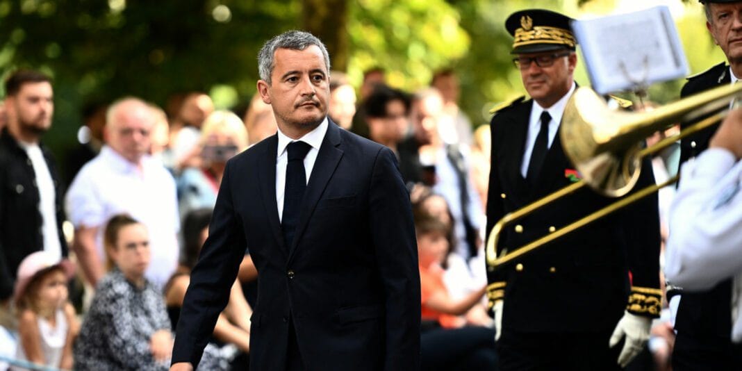 Gérald Darmanin en costume lors d'une cérémonie publique avec un orchestre militaire.