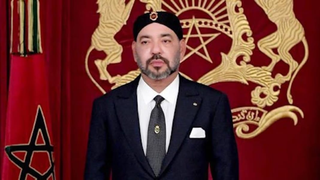 Fête de l’indépendance Marocaine : Un retour sur l’héroïsme d’un combat historique Homme en costume noir avec cravate devant drapeau marocain et symbole royal.