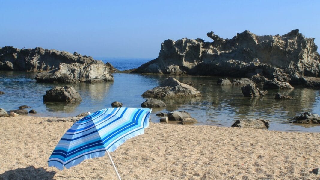 Touriste en Algérie : voici les codes à respecter pour éviter des problèmes Plage algérienne avec parasol bleu et rochers au bord de la mer.
