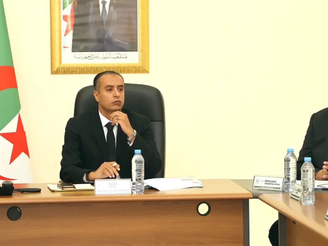 Walid Sadi en réunion, portant un costume noir, dans un bureau avec drapeau algérien en arrière-plan.