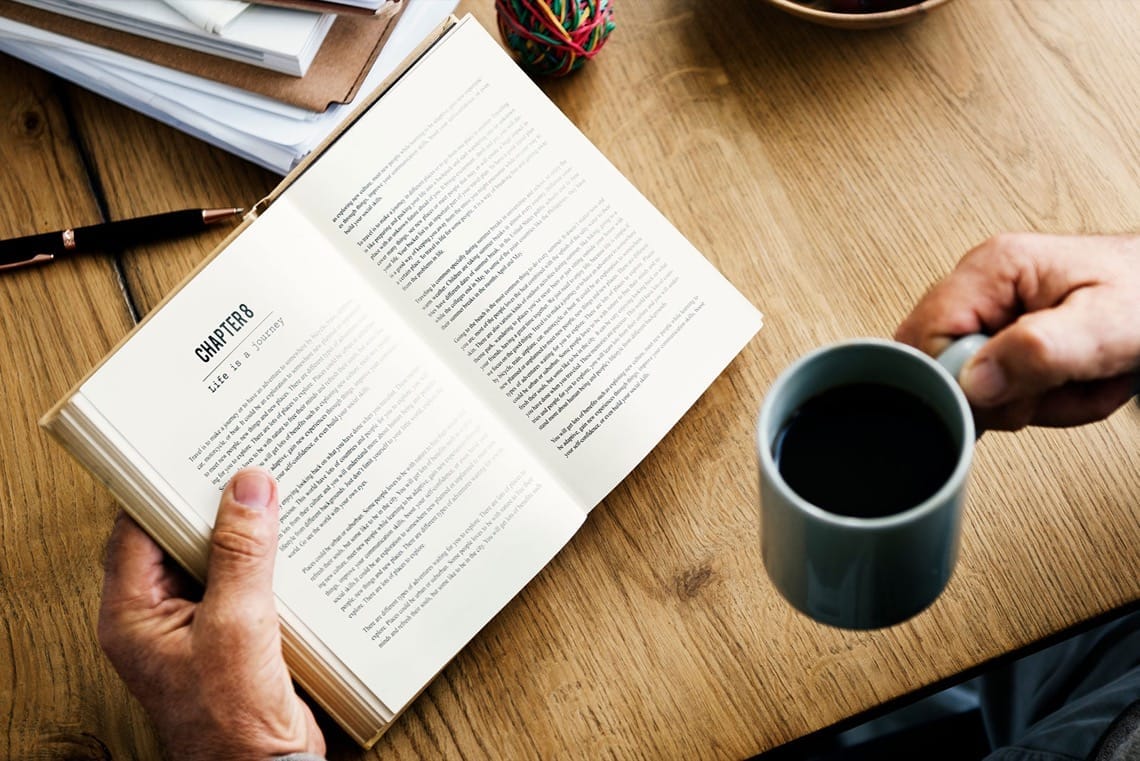 Personne lisant un livre avec une tasse de café sur un bureau en bois.
