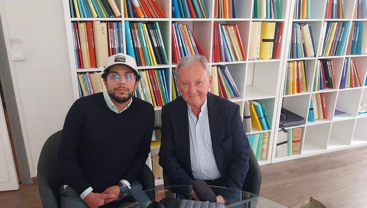 Deux hommes assis à une table en bibliothèque, livres en arrière-plan.
