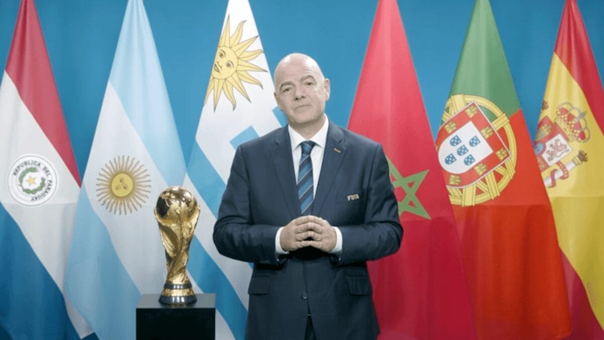 Homme en costume devant drapeaux de différents pays et trophée mondial.