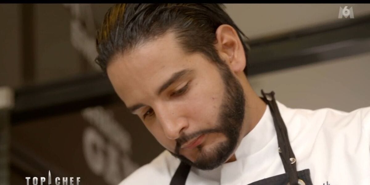 Chef concentré en cuisine, portant un uniforme blanc et un tablier noir.