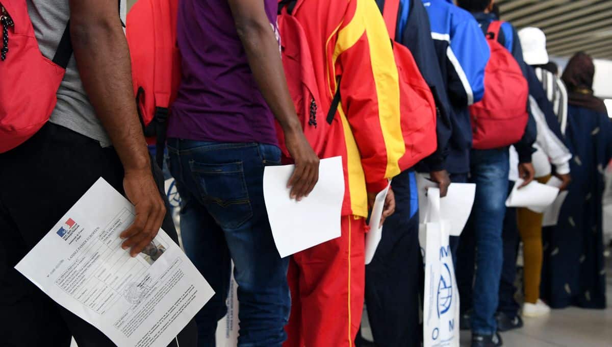 Demandeurs d'asile en file d'attente avec documents et sacs.