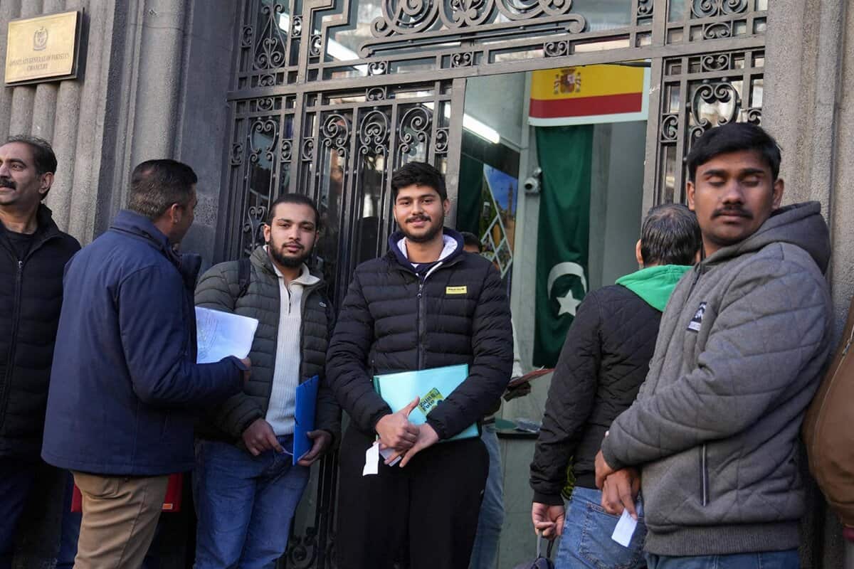 Groupe de migrants devant une ambassade avec drapeaux espagnol et pakistanais.