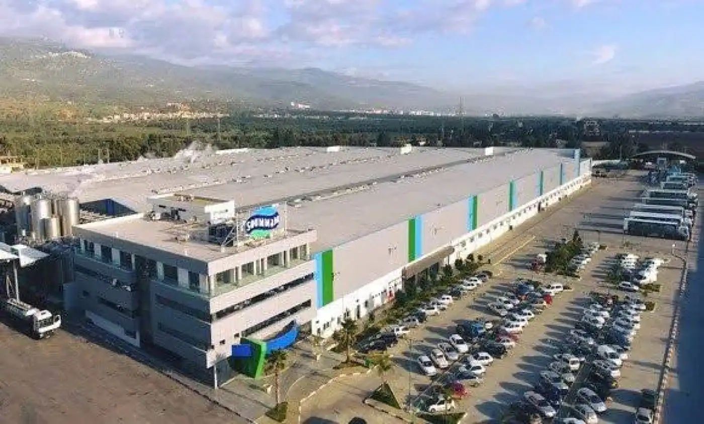 Grande usine industrielle avec parking spacieux et paysage montagneux en arrière-plan.