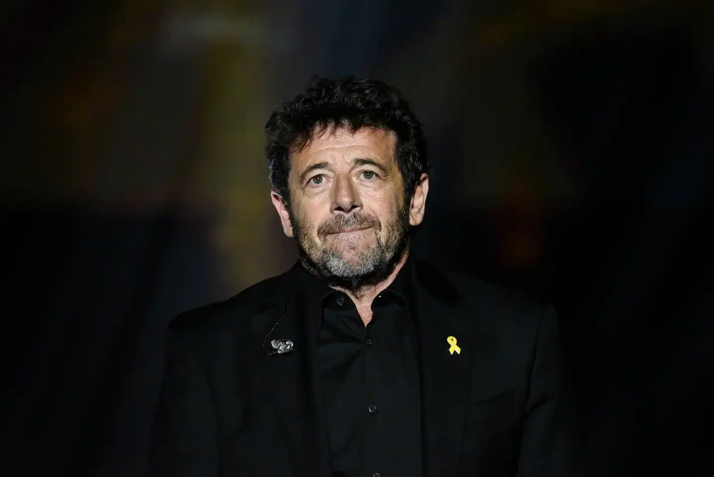Patrick Bruel portant un costume noir avec un ruban jaune.