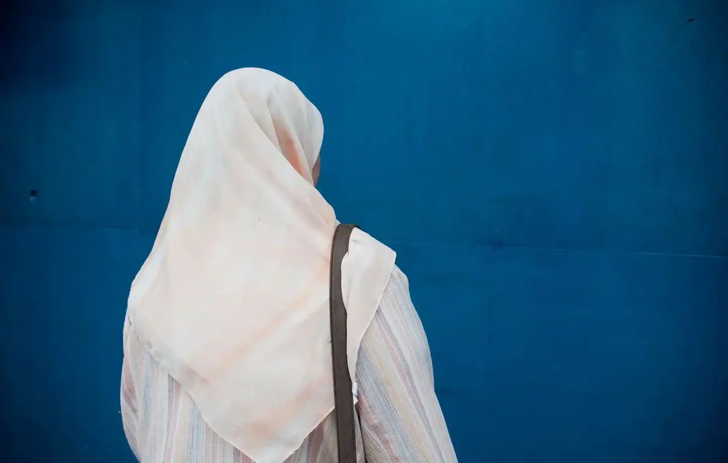 Femme avec hijab blanc vue de dos devant un mur bleu.