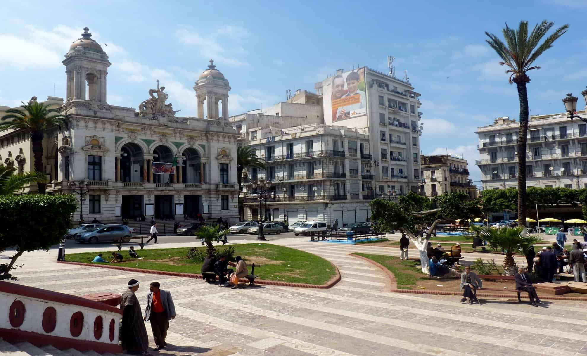 Vue d'une place animée avec bâtiments historiques et palmiers en Algérie.