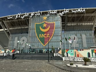 Stade Ali La Pointe à Douera avec drapeaux et logo MCA.