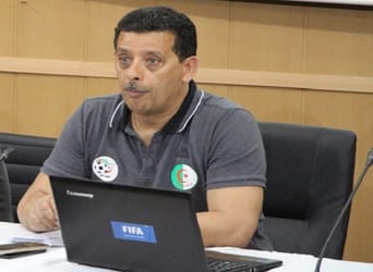 Taoufik Korichi en réunion, portant un polo gris avec logos FIFA et Algérie.