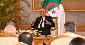 Président Tebboune parlant lors d'une réunion avec drapeau algérien en arrière-plan.