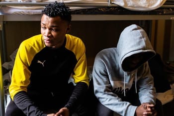 Deux jeunes hommes assis sous un lit, l'un en veste jaune et l'autre en sweat à capuche gris.