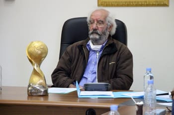Homme âgé avec barbe et lunettes, assis à un bureau avec trophée et documents.