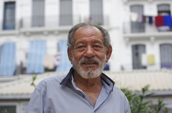 Homme âgé avec barbe souriant en extérieur urbain.