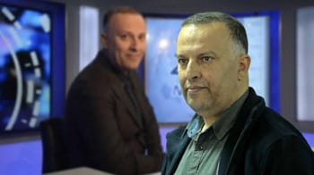 Deux hommes dans un studio de télévision avec écrans en arrière-plan.