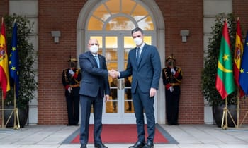 Deux hommes en costume se serrent la main devant un bâtiment officiel.