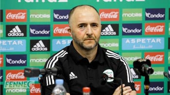 Entraîneur de football en conférence de presse avec fond de logos de sponsors.