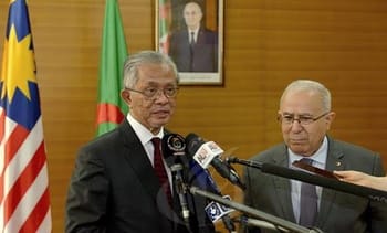 Deux hommes politiques lors d'une conférence en Algérie avec drapeaux et portrait en arrière-plan.