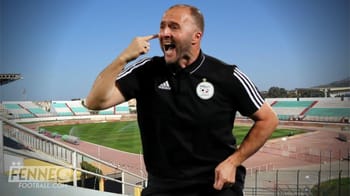 Djamel Belmadi souriant et pointant sa tête dans un stade vide.
