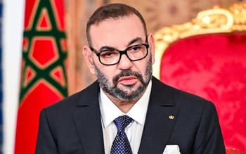 Homme en costume noir avec lunettes, siège dans un cadre officiel marocain.