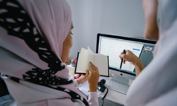 Deux femmes travaillent ensemble devant un ordinateur et un carnet.