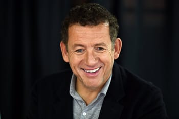 Dany Boon souriant lors d'une interview, portant un blazer noir et une chemise grise.