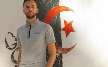 Jeune homme avec polo gris devant graffiti de la lune et étoile algérienne.