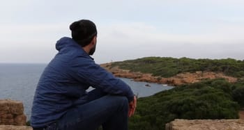 Homme assis contemplant la mer et la côte verdoyante.