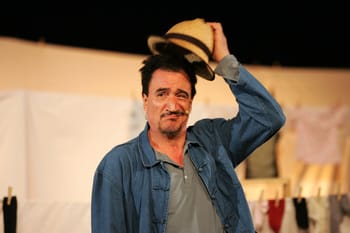 Comédien Fellag avec un chapeau, scène de théâtre en arrière-plan.