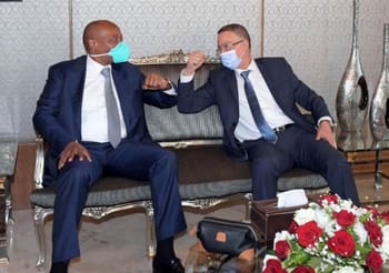 Deux hommes en costume échangent un salut avec un geste de coude, portant des masques faciaux.