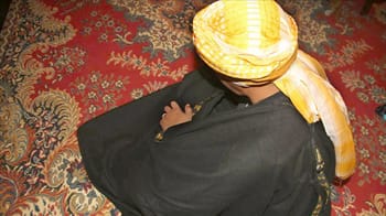 Homme portant un turban algérien traditionnel sur un tapis coloré.