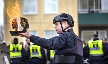 Policier en tenue de protection tenant un livre en flammes lors d'une manifestation.