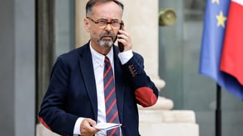 Robert Ménard parlant au téléphone devant un bâtiment officiel avec drapeaux européens et français.