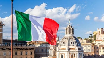 Drapeau italien flottant dans le ciel avec bâtiments historiques en arrière-plan.