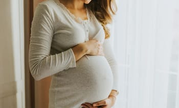 Femme enceinte tenant son ventre devant une fenêtre lumineuse.