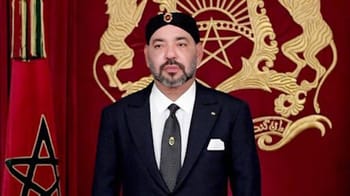 Homme en costume noir avec cravate devant drapeau marocain et symbole royal.